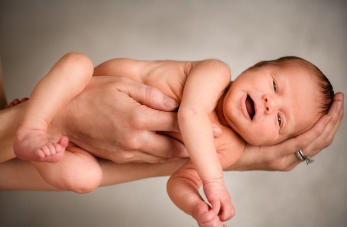 Recém-nascido sendo segurado por mãos adultas, com expressão tranquila, em fundo neutro, representando cuidados com a saúde infantil